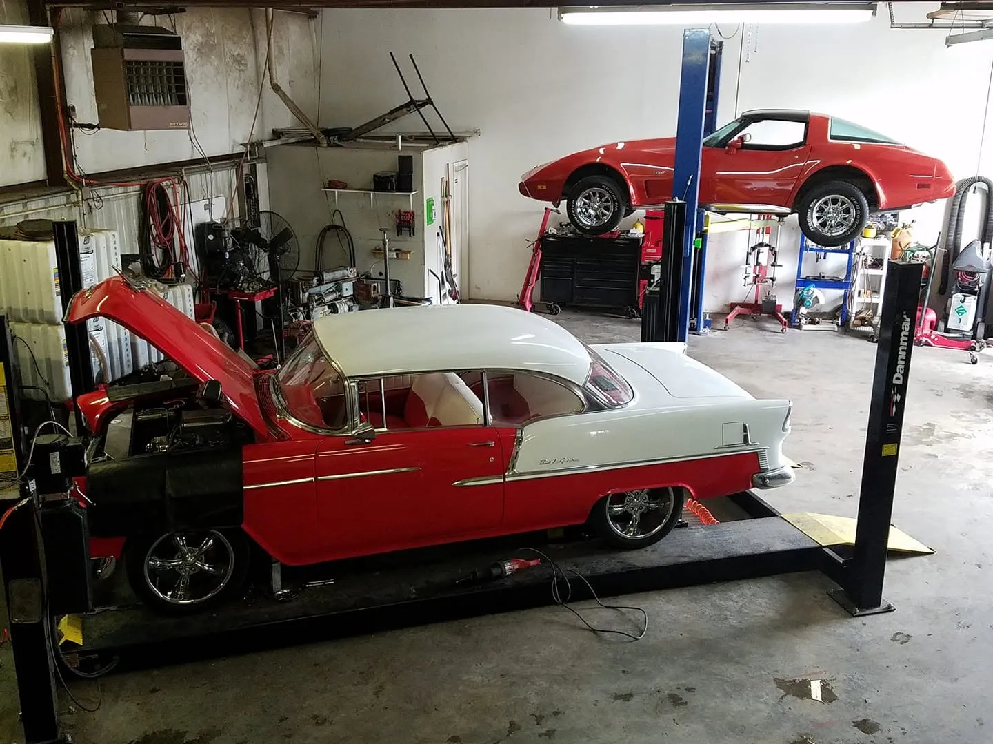 A red and white 1955 Chevrolet Bel Air with the hood up on a four-post lift, a red Corvette raised on the lift behind it