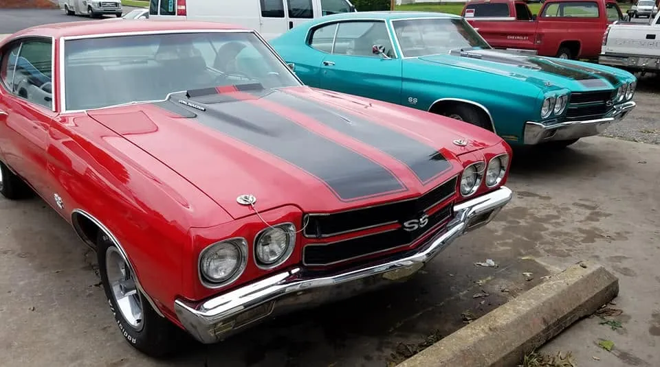 A red Chevelle SS with black stripes parked beside a turquoise Chevelle SS outside the shop